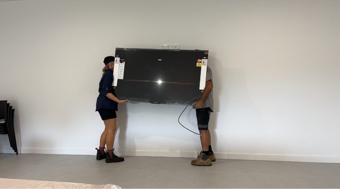Nic and Naomi from Orara TV Solutions lifting a large TV onto a wall mount